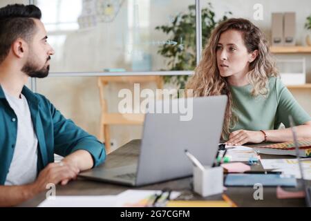 Young businesswoman sitting at the table and talking to her colleague they using laptop in their work at office Stock Photo