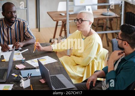 Female business leader in elegant dress talking to her employees and giving them instruction in work during a meeting at office Stock Photo