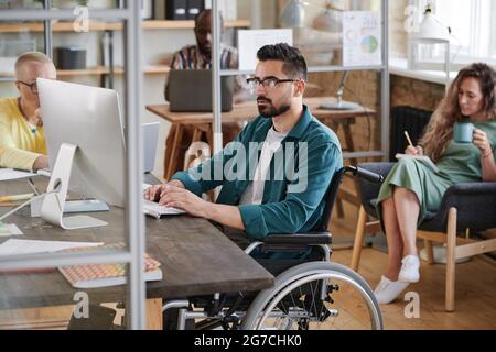 Serious young businessman in wheelchair sitting at the table and typing on computer he working at office with his colleagues Stock Photo