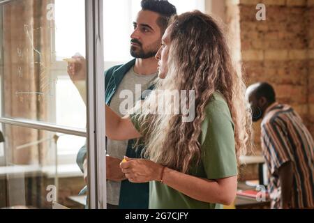 Two business colleagues drawing graphics together on glass wall and discussing it during presentation at office Stock Photo