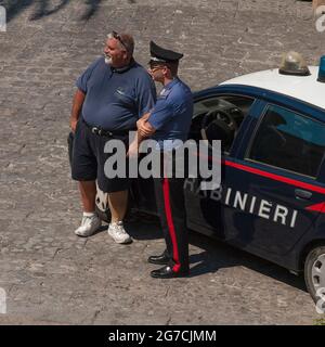 A young Carabiniere, an armed officer in Italy’s Arma dei carabinieri military police force, chats with a local resident leaning against his Fiat patrol car as he keeps a watchful eye on tourists walking in Via Seminario below the forecourt terrace of the Duomo di Cefalu, the historic cathedral in Cefalu, Sicily, Italy. Stock Photo