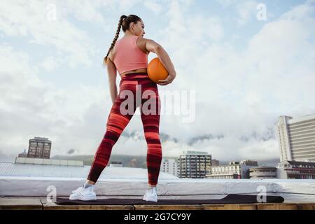 Rear view of woman in sportswear holding basketball standing outdoors. Athlete woman standing on rooftop with city seen in background. Stock Photo