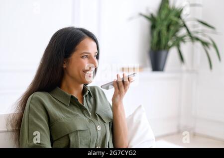 Charming indian woman is recording voice message and smiling, sits on the sofa at home and holding smartphone near mouth and talking in the mic, chatting, using voice recognizable mobile application Stock Photo