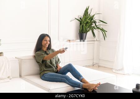 Full length of young Indian woman sits on the comfortable sofa at home and holds remote control, watching TV and resting alone, spends leisure time watching series Stock Photo