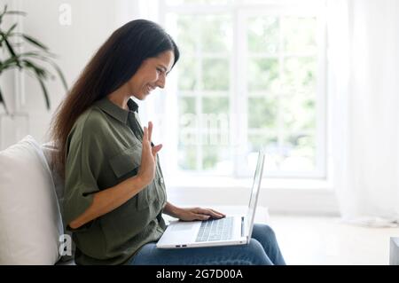 Positive smart young indian female freelancer or a student sitting on the couch with laptop on the lap, happy young eastern woman taking a part in virtual meeting, video calling and talking online, Stock Photo