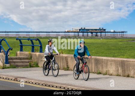 Southport, Merseyside.  Uk Weather. 13 July 2021: Early sunshine and warming temperatures reach 20C, as local residents take light exercise on the resort seafront promenade. Activities on the Coastal road as the dune habitat & grassing of the sands (Accretion), appears to extend across to the Fylde Coast.  The Sefton coast is struggling to repel two invaders – the sea and tough grasses sprouting over the beach which will eventually engul the pier as sand dunes form. Credit; MediaWorldImages/AlamyLiveNews Stock Photo