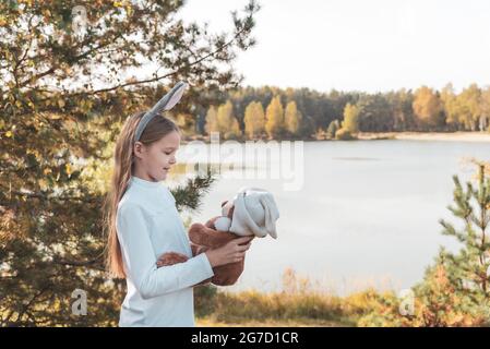 Happy girl with rabbit ears holds teddy bear on riverbank Stock Photo
