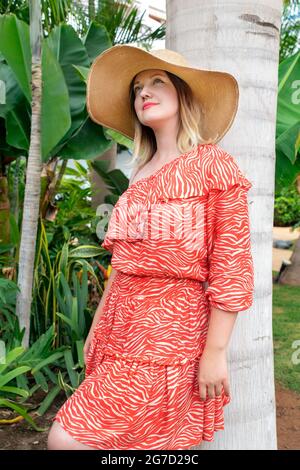 Dreamy and romantic portrait of a young attractive Caucasian millennial woman wearing a summer red dress, an oversized straw hat and leaning on a palm Stock Photo