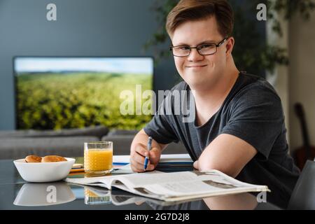 Young man with down syndrome smiling and doing home work in kitchen at home Stock Photo