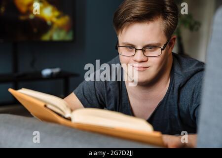 Young man with down syndrome reading book while lying on couch at home Stock Photo