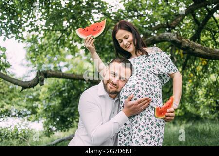 Handsome man listening to pregnant belly of his charming wife on fresh air. Pretty woman smiling sincerely and holding pieces of watermelon in hands. Stock Photo
