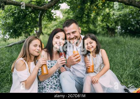 Portrait of caucasian parents with two cute daughters drinking juice in glass bottle from colorful straws. Smiling family spending cheerfully free time on nature. Stock Photo