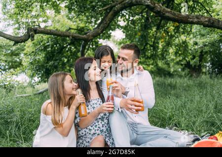 Portrait of caucasian parents with two cute daughters drinking juice in glass bottle from colorful straws. Smiling family spending cheerfully free time on nature. Stock Photo