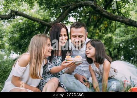 Caucasian father, mother and two little daughters eating homemade cupcakes during picnic at green garden. Happy family enjoying sweet dessert on fresh air. Stock Photo