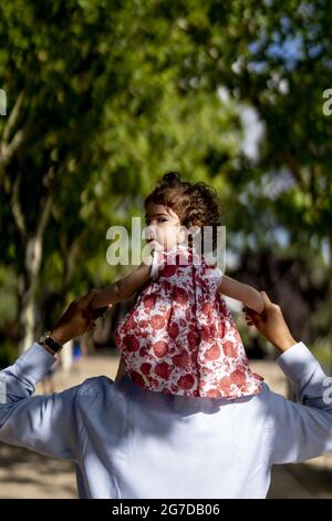 Vertical back view of the father carrying his daughter on his shoulders while in a park Stock Photo