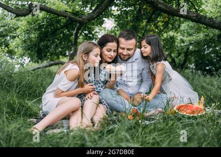 Caucasian father, mother and two little daughters eating homemade cupcakes during picnic at green garden. Happy family enjoying sweet dessert on fresh air. Stock Photo