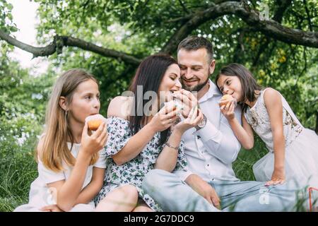 Positive parents with their little daughters sitting on green grass and tasting cupcakes. Happy family enjoying summer picnic on fresh air. Stock Photo