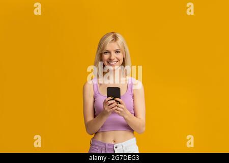Great app. Positive young woman holding and using cellphone, chatting online isolated on yellow studio background Stock Photo