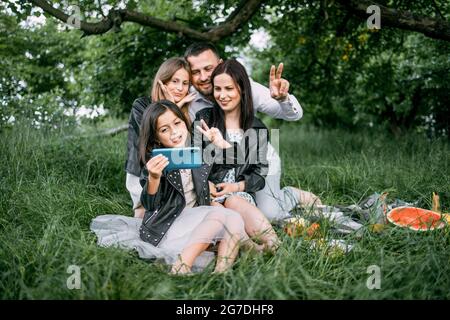 Cheerful parents and their cute daughters sitting together on green grass and using modern cell phone for taking self portrait. Concept of people, leisure and technology. Stock Photo