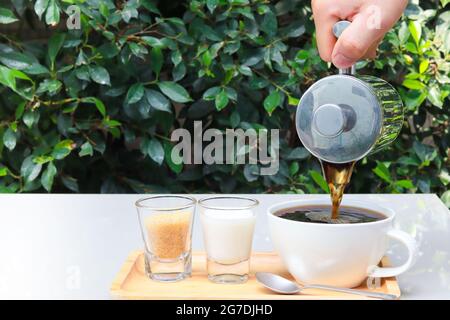 Pour coffee into a cup on a wooden tray with milk and sugar next to it. Tables are located in the restaurant, garden setting. Copy space. Stock Photo