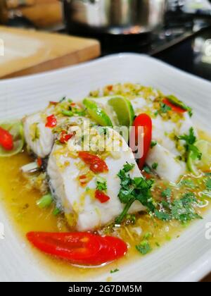 Thai food, steam fish lime sauce, served on white ceramic plate. Sliced lime, red chili pepper and chopped coriander on top. Kitchen atmosphere. Stock Photo