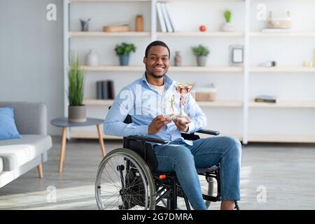 Happy paraplegic sportsman in wheelchair holding trophies and smiling at camera indoors Stock Photo