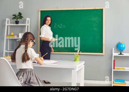 Primary school teacher at chalkboard and schoolgirl writing sitting at desk Stock Photo