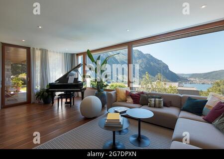 Living room with large light-colored sofa and lots of cushions and a black piano. Large window overlooking the valley with a lake view. Nobody inside Stock Photo