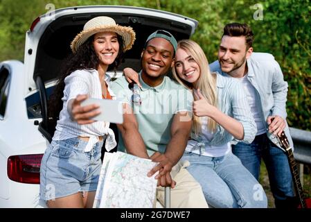 Funky young friends taking selfie on mobile phone during road trip, sitting in car trunk, traveling together in summer Stock Photo