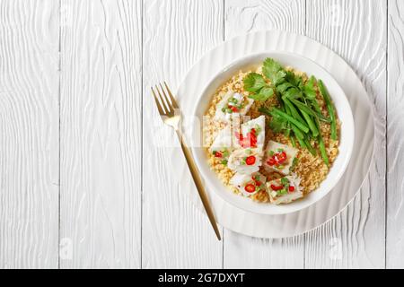 cod with bulgur, steamed green beans, sprinkled with hot chili peppers and spring onions in a white bowl, on a wooden table, balkan cuisine, flat lay, Stock Photo