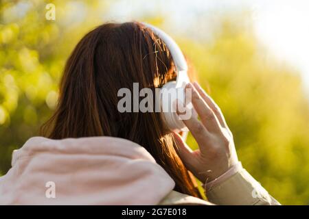 Back view of caucasian young woman listening music with headphones on sunny day while walking in city park Stock Photo