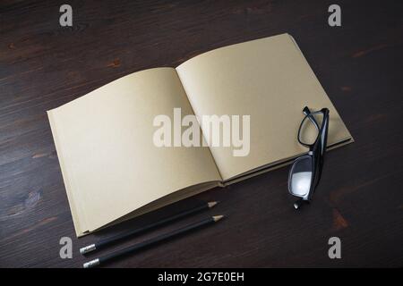 Opened book with blank craft paper pages, pencils and glasses on wooden background. Stock Photo