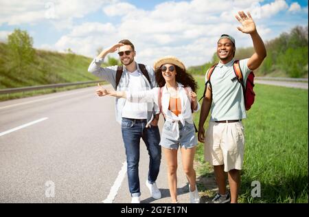 Millennial people standing on roadside, stopping car, trying to get free ride, having autostop journey, copy space Stock Photo