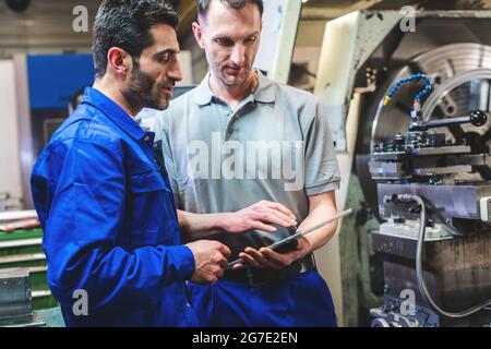 Two workers discussing a project in front of CNC lathe machine writing in clipboard Stock Photo