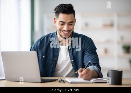 Joyful arab man taking notes while working with laptop Stock Photo