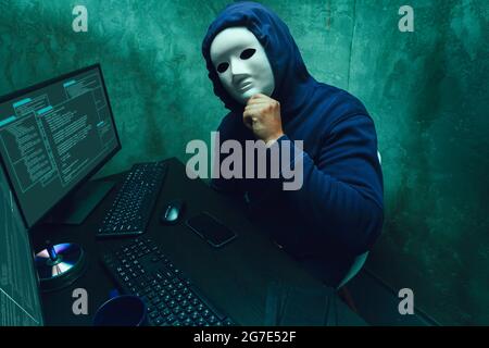 Anonymous hacker wearing face mask working on computer in dark room Stock Photo