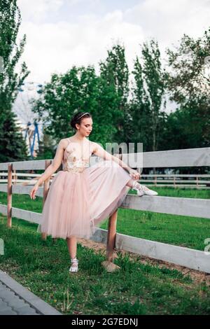 a young elegant ballerina has lifted her foot on the wooden board of the fence of the horse pen in the amusement park and is adjusting the satin
