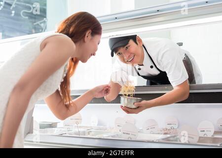 Helpful young sales assistant smiling at a customer through the glass of the display counter as he leans forwards to serve her selection of food Stock Photo