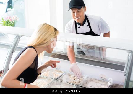 Friendly smiling deli worker helping a customer as she stands in front of a glass counter selecting food from trays Stock Photo