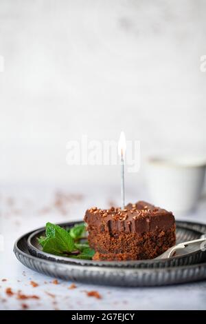 A Square of Chocolate cake topped with ganache, bronze sprinkles and a lit candle. A small piece has been remove from the square using the fork on the Stock Photo