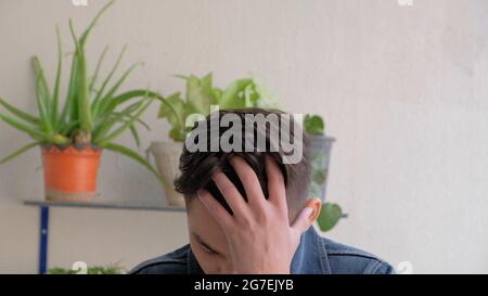Young depressed Indian male holding his head in his hand with houseplants in the background Stock Photo