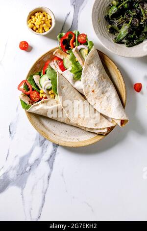 Traditional mexican tortila wrap with pork meat and vegetables in ceramic plate served with grilled green peppers jalapenos and corn over white marble Stock Photo