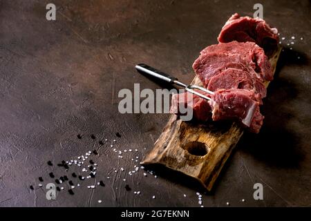 sliced raw beef tenderloin meat for steaks on wooden board with metal meat fork, salt and pepper over dark brown texture background. Food cooking back Stock Photo