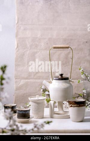 White and black craft handmade ceramic teapot with black ceramic mugs, katakuchi jug and blossoming cherry tree as decoration standing on marble table Stock Photo