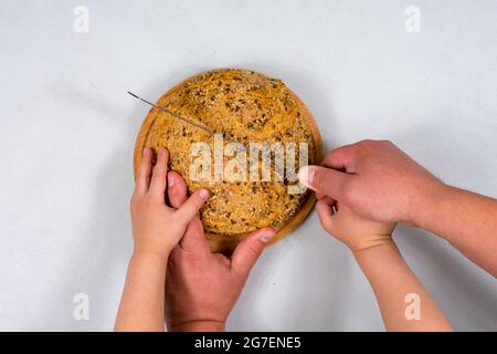 With two hands, men and children cut wheat bread on a wooden board. Teaching children. Homemade bread with grains. Stock Photo