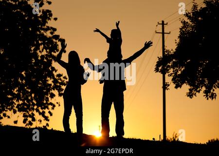 Family having a walk at sunset, the child sitting on his father's shoulders waving at the viewer; the whole scene is shot back lit, very tranquil and Stock Photo