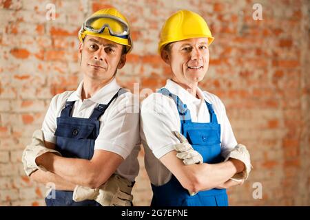 Two construction workers standing in a construction site in front of a war brick wall. Their arms are folded and they are wearing hardhats Stock Photo