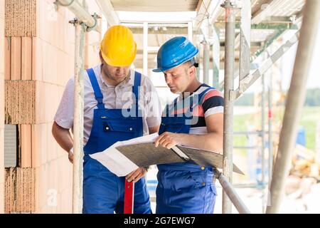 Two construction workers on site with helmets having discussion in scaffold with plans or blueprints Stock Photo