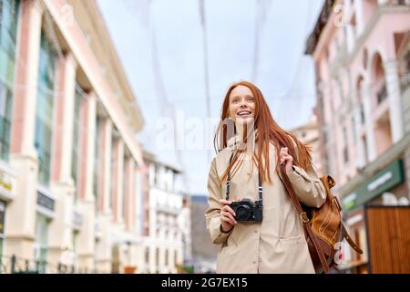 Young smiling woman tourist walks around city in spring, holds retro camera. Redhead female on trip, in casual urban style outfit. Portrait of caucasi Stock Photo