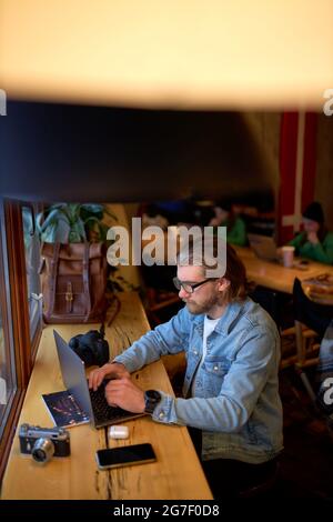 Young caucasian male photographer working at desktop with laptop computer using wireless internet connection in coffee shop.Young hipster man sitting Stock Photo